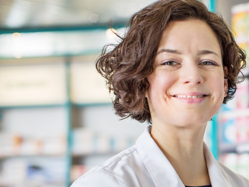Gut gelaunte Apothekerin vor einem Regal mit Medikamenten und Drogeriebedarf